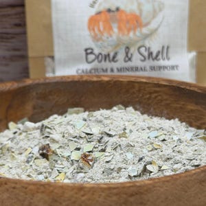 May include: A wooden bowl filled with a coarse, light-colored powder containing small, fragmented pieces of shell. Behind the bowl, a tan paper package displays a hermit crab illustration and the text Bone & Shell CALCIUM & MINERAL SUPPORT.
