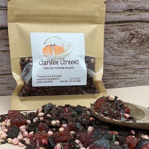 May include: A brown paper pouch labeled Hermit's Pantry Garden Greens Veggie Power Blend, Net Wt. 0.5 oz (14g), 2.7 x 3.5 inches. The pouch contains a dark red, green, and white dried vegetable mix, with a wooden spoon holding a portion of the blend.