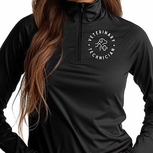 Op de afbeelding: Zwart sportshirt met lange mouwen en een kraag met een kwart rits. De shirt heeft een witte cirkelvormige afbeelding op de borst met de woorden "VETERINARY TECHNICIAN" rond een hond, kruis en medisch symbool.