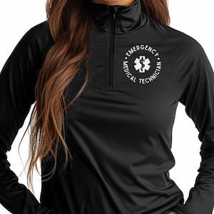 Pode incluir: Camisa preta de manga comprida com gola com zíper. A camisa apresenta um emblema branco com as palavras "EMERGENCY MEDICAL TECHNICIAN" circundando um símbolo médico branco. A camisa é feita de um tecido escuro e liso.