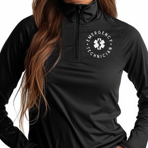 May include: Black long-sleeve shirt with a quarter-zip collar. The shirt features a white circular emblem with a medical symbol and the text "EMERGENCY TECHNICIAN" in a circular pattern around it. The shirt is made of a smooth, possibly athletic material.