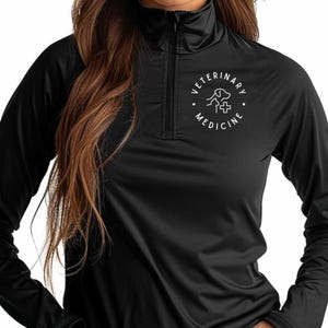 May include: Black long-sleeve athletic shirt with a quarter-zip collar. The shirt features a white circular logo on the chest with the words "VETERINARY MEDICINE" around a dog and cross graphic. The shirt is made of a smooth, likely moisture-wicking material.