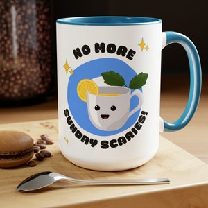 Op de afbeelding: Witte keramische mok met een blauwe rand en handvat. De mok heeft een cartoonillustratie van een theekopje met een citroenschijfje en een takje munt. De tekst "NO MORE SUNDAY SCARIES!" staat rond het theekopje.
