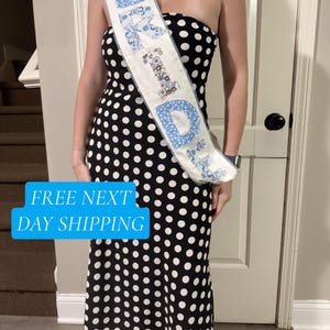 May include: A strapless black midi dress with a white polka dot pattern worn with a white sash featuring the blue floral patterned letters spelling BRIDE. The sash has a decorative lace trim along the edges and is worn diagonally across the torso.
