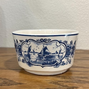 May include: A small, off-white ceramic bowl with a blue nautical scene. The bowl features a detailed illustration of sailboats, windmills, and water, framed by floral and scrollwork designs. The rim is trimmed with a blue border.