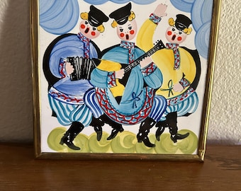 May include: Square ceramic tile with a gold frame featuring a folk art illustration of three musicians. The musicians are dressed in blue and yellow outfits, playing an accordion, guitar, and singing. The background has blue cloud-like shapes.