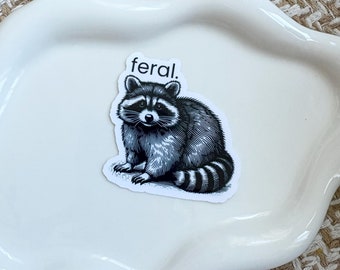 May include: A white ceramic dish with a black and white sticker of a raccoon with the word "feral" above it.