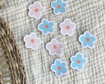 May include: Nine pastel flower stickers in blue and pink. The stickers are arranged in a random pattern on a white fabric background.