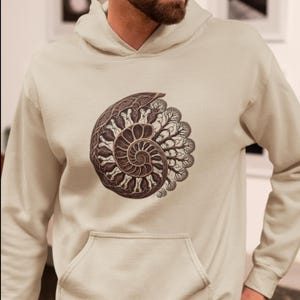 May include: A beige hooded sweatshirt featuring a central graphic print of a dark brown ammonite fossil shell. The shell design incorporates intricate, swirling patterns and tree-like branch structures radiating from the center spiral.