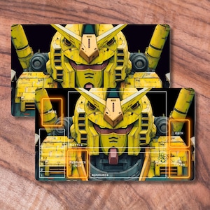 May include: Two yellow and grey playmats with a detailed illustration of a robot's face. The playmats have gameplay zones, including "Base," "Deck," and "Resource." The design is set against a black background.