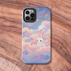 Op de afbeelding: Een telefoonhoesje met een pastelblauwe, roze en paarse wolkenachtergrond met een wit konijn dat op een wolk zit. Het hoesje heeft een zwarte camera lens uitsparing en een sterrenhemel ontwerp.