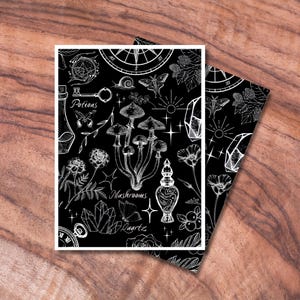 May include: Two black and white art prints with botanical and mystical illustrations. The designs include mushrooms, crystals, potions, and a compass. The word "Mushrooms" is visible on one print. The prints are displayed on a wooden surface.