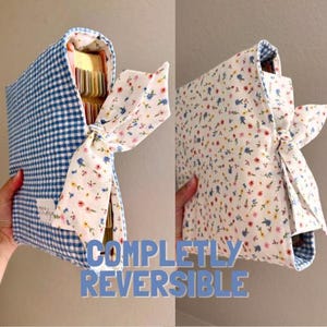 May include: A reversible fabric book cover featuring a blue and white gingham pattern on one side and a white floral print on the other. The cover includes a fabric tie closure and a small white rectangular label with text on the gingham side.
