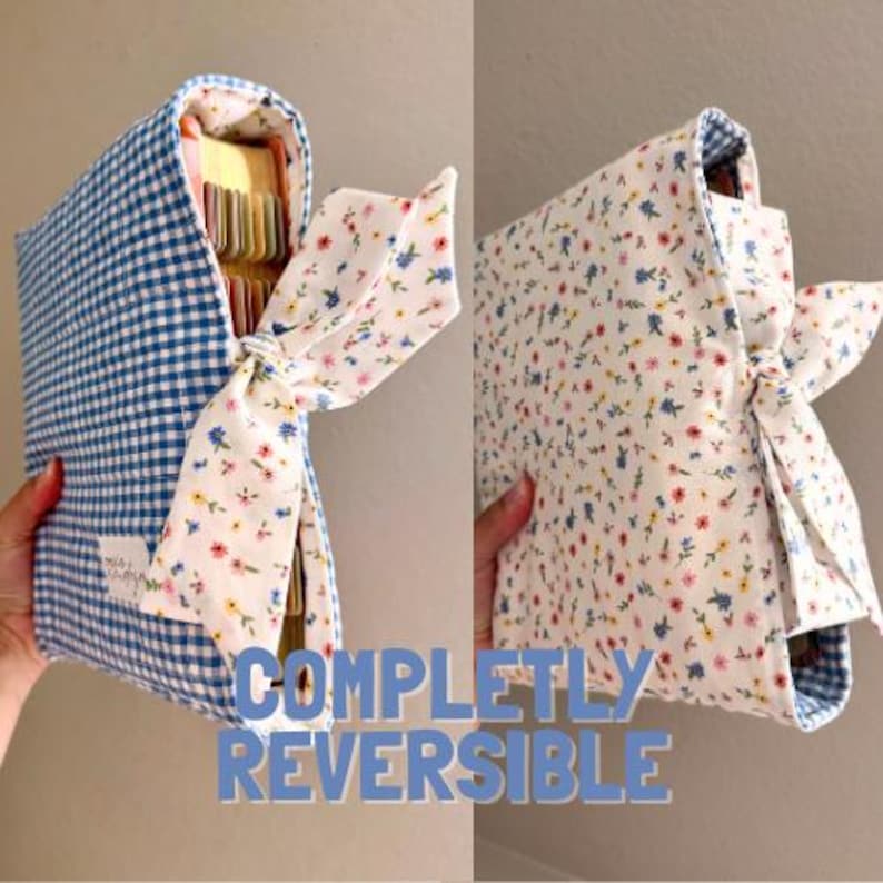 May include: A reversible fabric book cover featuring a blue and white gingham pattern on one side and a white floral print on the other. The cover includes a fabric tie closure and a small white rectangular label with text on the gingham side.