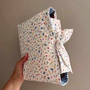 May include: A fabric book cover featuring a white background with a small-scale floral print in red, yellow, and blue. The cover includes a side tie closure made of the same floral fabric and is lined with a blue and white gingham checkered pattern.