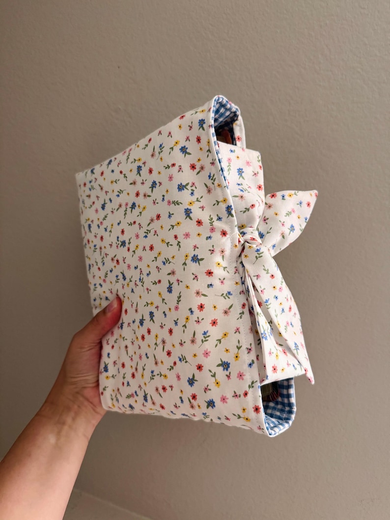 May include: A fabric book cover featuring a white background with a small-scale floral print in red, yellow, and blue. The cover includes a side tie closure made of the same floral fabric and is lined with a blue and white gingham checkered pattern.