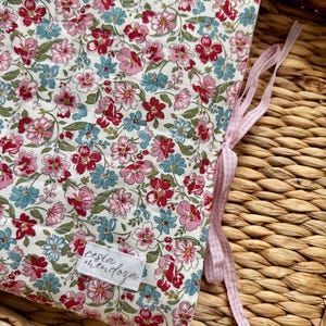 May include: A quilted fabric cover featuring a floral pattern with pink, red, and light blue flowers on a white background. The cover includes a white rectangular label with the text cesia mendoza and light pink gingham ribbon ties.