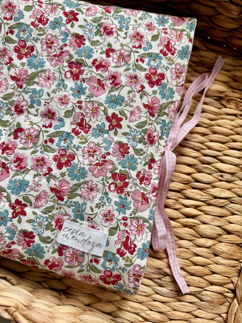 May include: A quilted fabric cover featuring a floral pattern with pink, red, and light blue flowers on a white background. The cover includes a white rectangular label with the text cesia mendoza and light pink gingham ribbon ties.