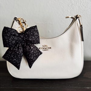 May include: A white leather hobo bag with a gold-tone zipper closure and the Coach New York logo in gold lettering. A large black fabric bow with a small grey floral pattern is attached to the side of the bag with a gold-tone metal clip and ring.