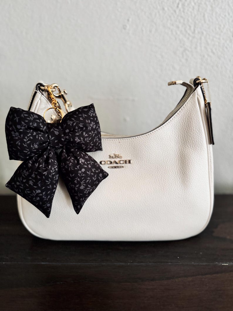 May include: A white leather hobo bag with a gold-tone zipper closure and the Coach New York logo in gold lettering. A large black fabric bow with a small grey floral pattern is attached to the side of the bag with a gold-tone metal clip and ring.