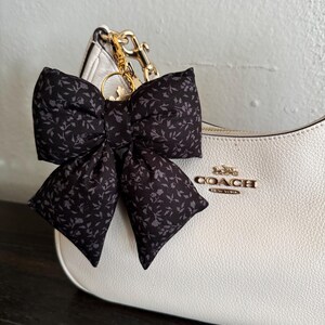 May include: A black fabric bow with a small gray floral pattern attached to a white leather handbag by a gold-toned metal clip. The handbag features the gold-toned text COACH NEW YORK on the front, with a textured surface and a curved top edge.