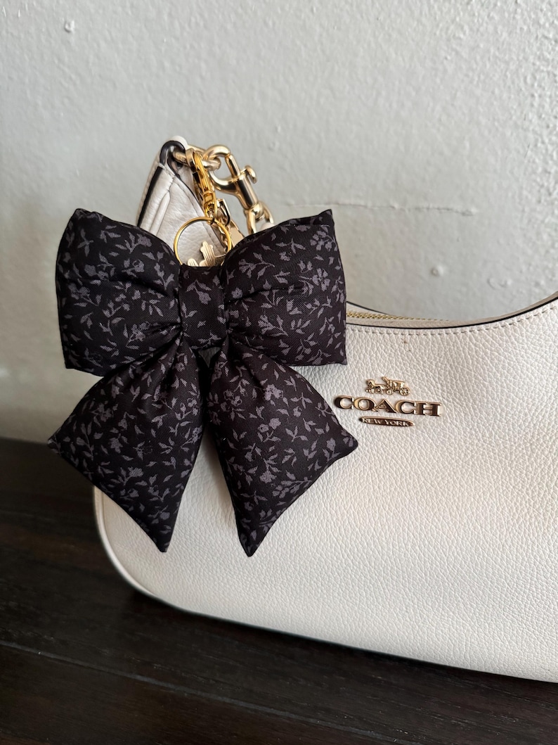 May include: A black fabric bow with a small gray floral pattern attached to a white leather handbag by a gold-toned metal clip. The handbag features the gold-toned text COACH NEW YORK on the front, with a textured surface and a curved top edge.