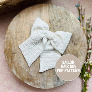 May include: A white hair bow with a textured, crinkled fabric surface. The bow features a central knot and two long, wide tails extending downward. A circular beige graphic overlay on the lower right displays the text SAILOR HAIR BOW PDF PATTERN.