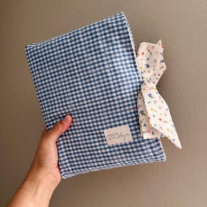 May include: A rectangular fabric cover featuring a blue and white gingham pattern. A white fabric label with the text cesia mendoza is stitched on the front. A white fabric tie with a small multicolored floral print is attached to the side.