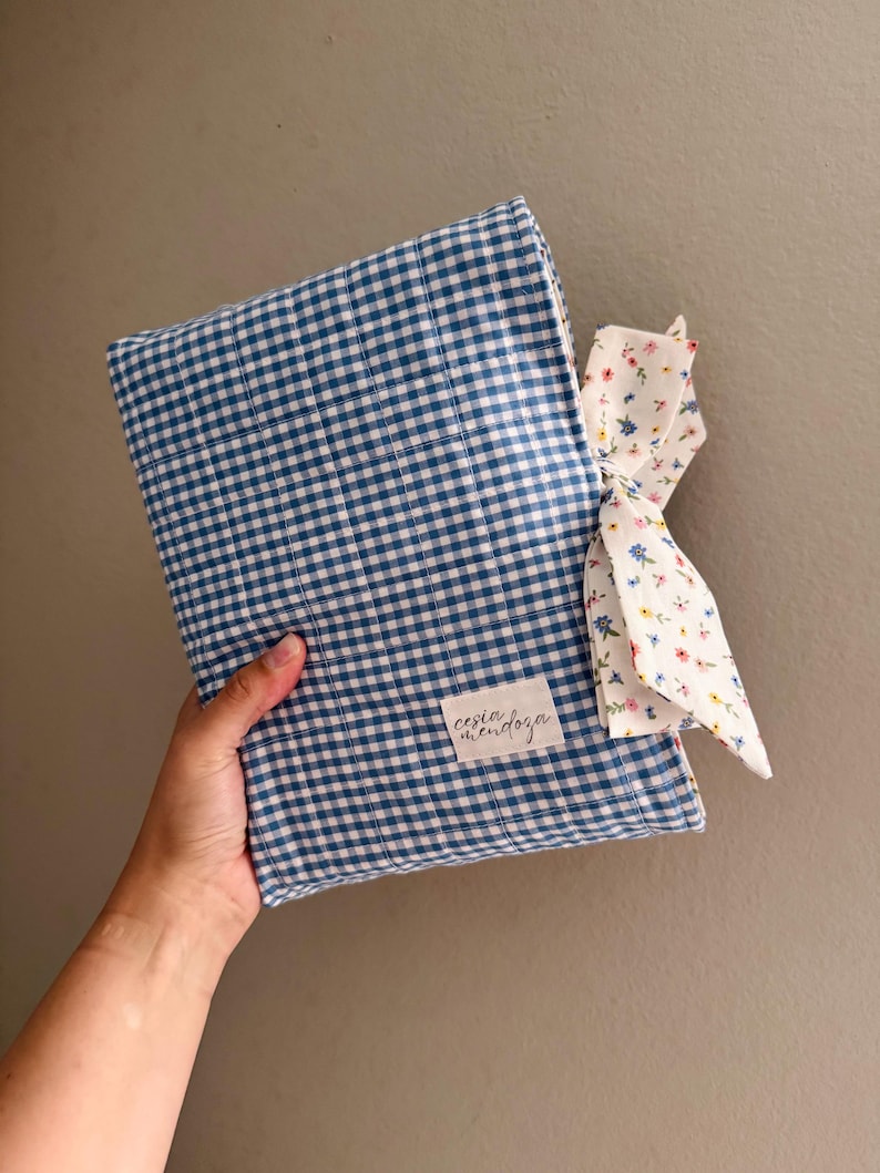 May include: A rectangular fabric cover featuring a blue and white gingham pattern. A white fabric label with the text cesia mendoza is stitched on the front. A white fabric tie with a small multicolored floral print is attached to the side.