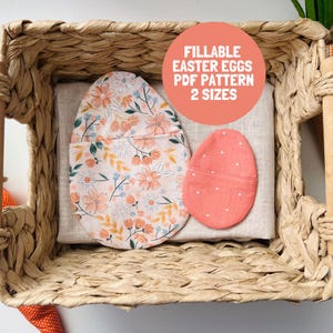 May include: Two fabric egg-shaped pouches resting on a light beige cloth inside a woven basket. One pouch features a floral pattern with peach, blue, and green tones, while the smaller pouch is solid coral with white dots. Text reads: FILLABLE EASTER EGGS PDF PATTERN 2 SIZES.
