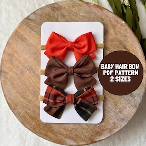 May include: Three fabric hair bows attached to a white display card. The top bow is solid orange, the middle bow is solid brown, and the bottom bow features a brown, orange, and black plaid pattern. Text on the card reads BABY HAIR BOW PDF PATTERN 2 SIZES.