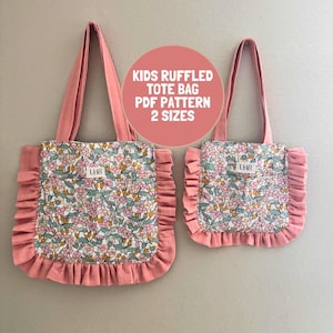 Pode incluir: Duas bolsas tote de tamanhos diferentes com um padrão floral de flores cor-de-rosa, verdes e amarelas sobre um fundo branco, acentuadas por bordas e alças com folhos em rosa sólido. Uma etiqueta branca com o texto LHB está centrada.