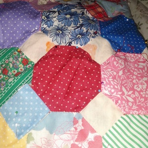 May include: A patchwork quilt in progress, featuring a variety of floral and geometric patterns in shades of red, blue, green, pink, and yellow. The quilt is pinned together with safety pins and is being sewn by hand.