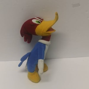 May include: A plush toy of Woody Woodpecker, a cartoon character. The toy is red, yellow, and blue. The toy is standing on a white surface.