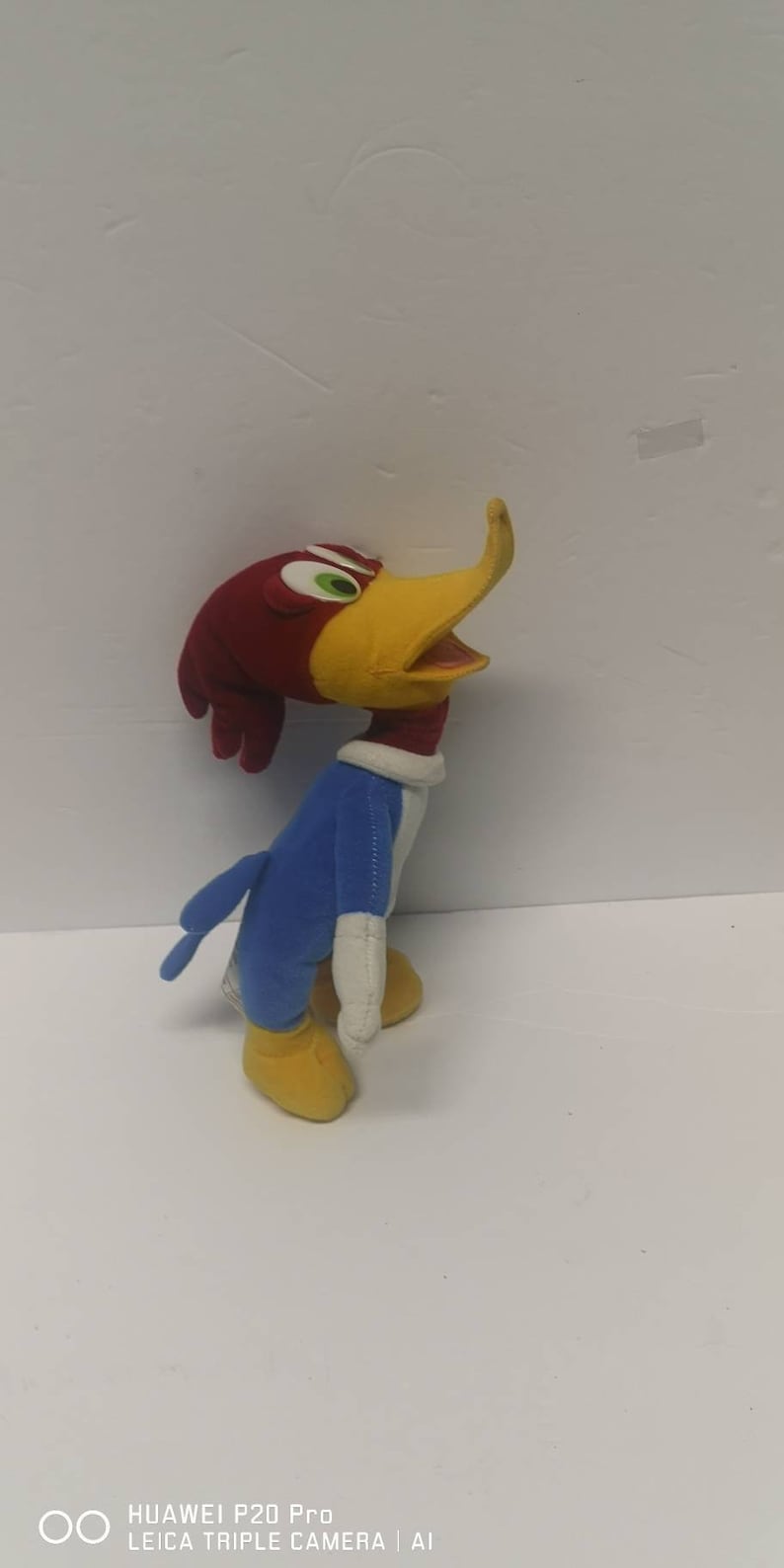 May include: A plush toy of Woody Woodpecker, a cartoon character. The toy is red, yellow, and blue. The toy is standing on a white surface.