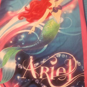 Puede incluir: Una manta con Ariel de La Sirenita de Disney. El dise&ntilde;o muestra a Ariel con cabello rojo ondeante, una cola verde y un top de concha p&uacute;rpura, sobre un fondo azul y rosa. El nombre "Ariel" est&aacute; escrito en una fuente estilizada.