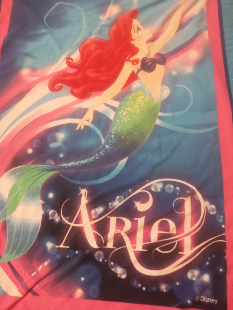 Puede incluir: Una manta con Ariel de La Sirenita de Disney. El dise&ntilde;o muestra a Ariel con cabello rojo ondeante, una cola verde y un top de concha p&uacute;rpura, sobre un fondo azul y rosa. El nombre "Ariel" est&aacute; escrito en una fuente estilizada.