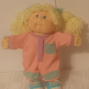 Peut inclure: Une poupée aux cheveux en laine blonde coiffés en couettes, aux yeux bleus et au visage rond. La poupée porte une combinaison rose à manches longues avec une bande centrale violette, une poche poitrine turquoise, des genouillères turquoise.