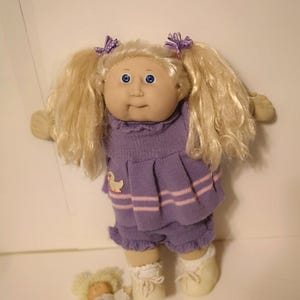 Peut inclure: Deux poupées Cabbage Patch Kids. La plus grande poupée a les cheveux blonds, les yeux bleus et porte une robe violette et rose avec un motif de canard. La plus petite poupée porte une chemise blanche avec des cœurs violets et se tient sur une surface blanche.