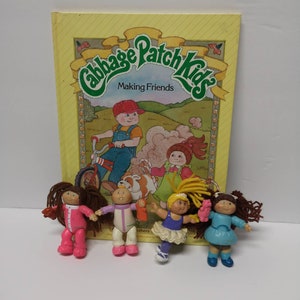 Peut inclure: Cinq poupées Cabbage Patch Kids, chacune avec une tenue et une coiffure différentes. Les poupées sont debout devant un livre intitulé "Cabbage Patch Kids Making Friends".