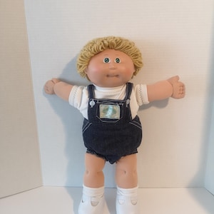 May include: A vintage doll with blonde yarn hair, blue eyes, and a rosy complexion. The doll is dressed in a white shirt, dark blue denim dungarees, white socks, and white shoes. The doll's arms are outstretched.