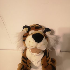 May include: A plush tiger toy with brown and black stripes, white fur on its chest and face, and a black nose. The tiger is sitting with its legs crossed and has a friendly expression.