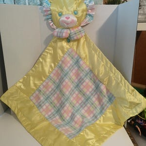May include: A yellow baby security blanket with a lion head and a plaid center. The lion has a pastel mane, blue eyes, and a pink nose. The blanket has a satin border.