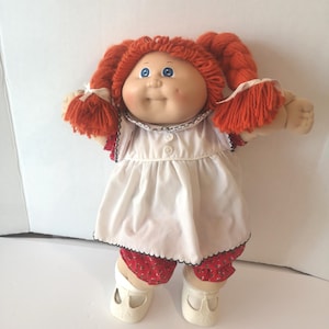 May include: A soft-sculpture doll with bright orange yarn hair styled in two pigtails, blue eyes, and dimpled cheeks. The doll wears a white sleeveless dress with a black scalloped trim, red floral-print bloomers, and cream-coloured plastic shoes.