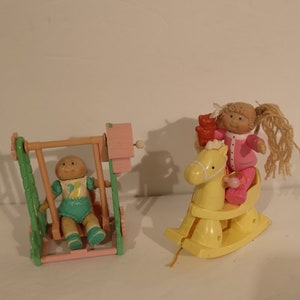 May include: Two Cabbage Patch Kids dolls, one on a pink and green swing set and the other on a yellow rocking horse. Both dolls are wearing pajamas.