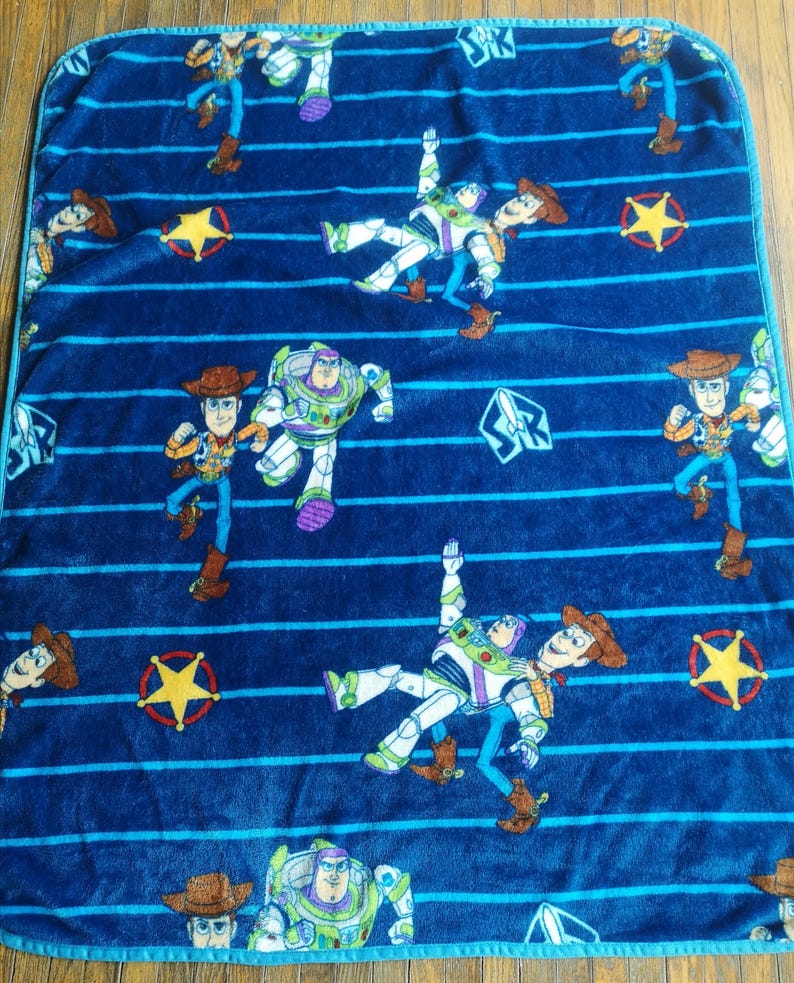May include: Blue fleece blanket with a repeating pattern of Woody and Buzz Lightyear from Toy Story. The blanket features blue and white stripes and yellow stars.