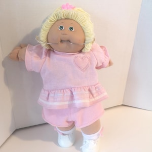 May include: A vintage doll with a round face, blonde yarn hair, and blue eyes. The doll wears a pink outfit with a heart on the chest, matching shorts, white shoes, and a metal dental appliance. A pink flower adorns the hair.