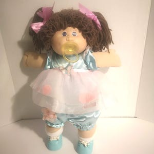 May include: A doll with brown yarn hair styled in pigtails with pink ribbons, wearing a light blue satin outfit with a white tulle overlay featuring pink polka dots and a butterfly accent. The doll has a yellow pacifier and light blue shoes.
