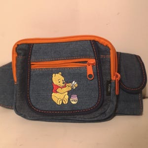 May include: Denim fanny pack with orange trim and zippers. Features an embroidered image of Winnie the Pooh holding flowers and a honey pot. The bag has a main compartment and a smaller front pocket.