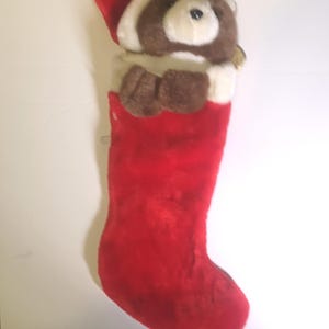 May include: A red Christmas stocking with a teddy bear face peeking out. The bear has brown fur, white accents, and a red Santa hat. A gold bell hangs from the stocking.